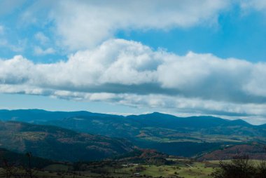 Yeşil, kırmızı, turuncu ve sarı renkli ormanlarla kaplı dağ manzarası manzaraya zıt düşüyor. Mavi ve gri gökyüzü ile doğanın güzel manzarası.