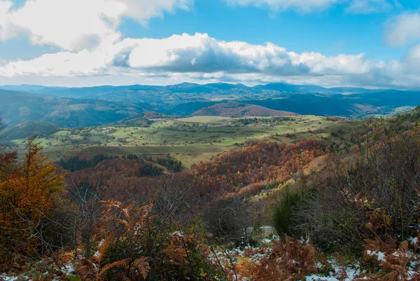 Yeşil, kırmızı, turuncu ve sarı renkli ormanlarla kaplı dağ manzarası manzaraya zıt düşüyor. Mavi ve gri gökyüzü ile doğanın güzel manzarası.
