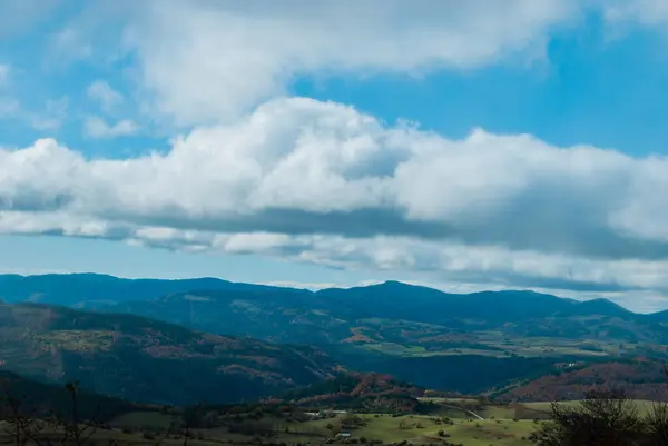 Yeşil, kırmızı, turuncu ve sarı renkli ormanlarla kaplı dağ manzarası manzaraya zıt düşüyor. Mavi ve gri gökyüzü ile doğanın güzel manzarası.