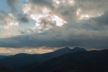 Dağlık arazi, bulutlu bir gün batımı gökyüzü, fırtınalı hava, dağlar yeşil ve turuncu renklerle ağaçlandırılmış..