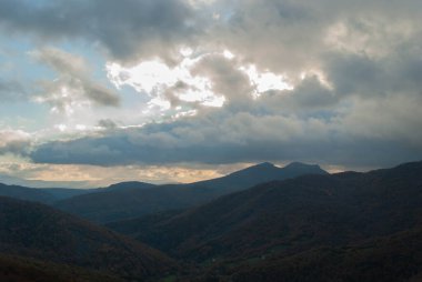 Dağlık arazi, bulutlu bir gün batımı gökyüzü, fırtınalı hava, dağlar yeşil ve turuncu renklerle ağaçlandırılmış..