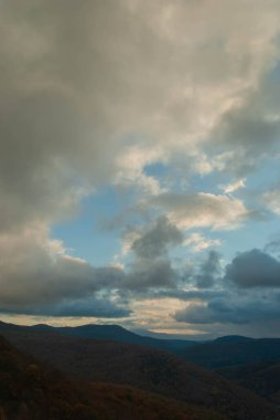 Dağlık arazi, bulutlu bir gün batımı gökyüzü, fırtınalı hava, dağlar yeşil ve turuncu renklerle ağaçlandırılmış..