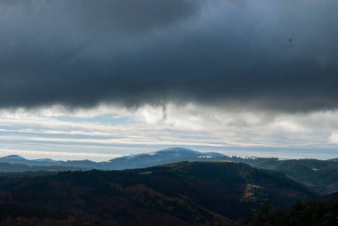 Dağlık arazi, bulutlu bir gün batımı gökyüzü, fırtınalı hava, dağlar yeşil ve turuncu renklerle ağaçlandırılmış..