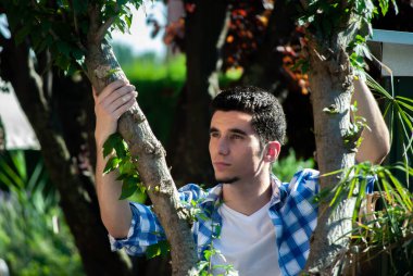 Genç adam yeşil bir bahçede iki ağaç arasında eğiliyor.