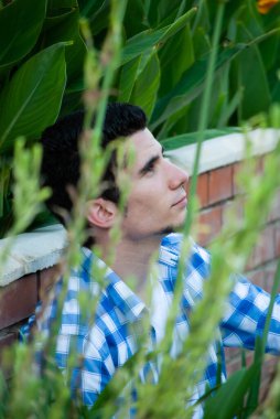 Önünde ve arkasında saksı bitkileri olan tuğlalara yaslanan genç bir adamın profili.