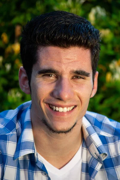 Kameraya gülümseyen genç bir adamın yakın plan portresi, arkasında bitkilerin yeşil bir arka planı var.