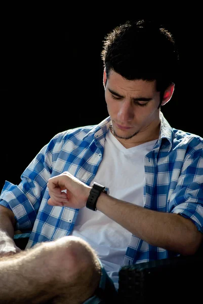 Kol saatine bakan zarif genç bir adam, arkasında koyu bir arka planla oturduğu yerde oturuyor.