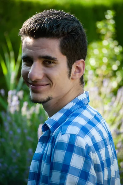 Genç adam yeşil bir ortamda kameraya gülümsüyor ve yan bakıyor, arkasında bitkilerin yeşil bir arka planı var.