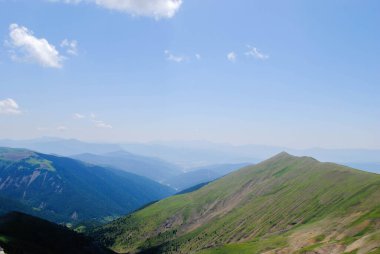 Dağlık yeşil dağ sıraları, açık mavi gökyüzü ve birkaç bulutla kaplı..