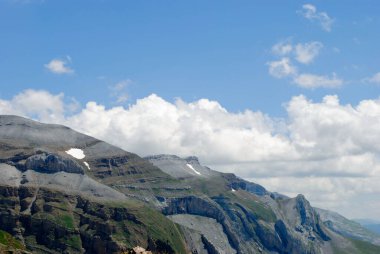 Dağlık yeşil dağ sıraları, açık mavi gökyüzü ve birkaç bulutla kaplı..