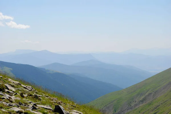 Dağlık yeşil dağ sıraları, açık mavi gökyüzü ve birkaç bulutla kaplı..