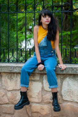 Sarı giyinmiş ve kot pantolon giymiş genç bir kız sonbahar yeşili ve sarı renkleri olan bir parkta taş bir duvarda oturuyor..