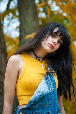 Sarı giyinmiş ve kot pantolon giymiş genç bir kız sonbahar yeşili ve sarı renkleri olan bir parkta poz veriyor..