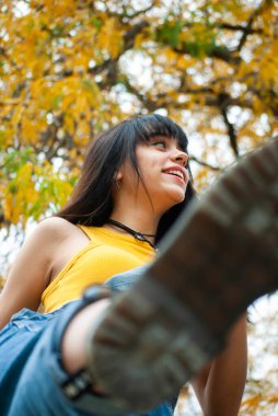 Sarı giyinmiş ve kot tulumlu genç bir kız sarı ve yeşil sonbahar dallarını ve yapraklarını gösteren yakın plan bir çekimde öne doğru tekmeliyor..