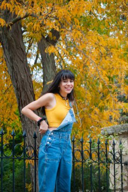 Sarı giyinmiş ve kot pantolon giymiş genç bir kız sonbahar yeşili ve sarı renkleri olan bir parkta gülümsüyor..