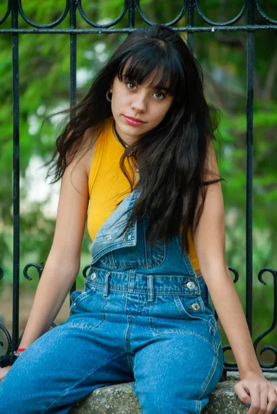 Sarı giyinmiş ve kot pantolon giymiş genç bir kız sonbahar yeşili ve sarı renkleri olan bir parkta taş bir duvarda oturuyor..