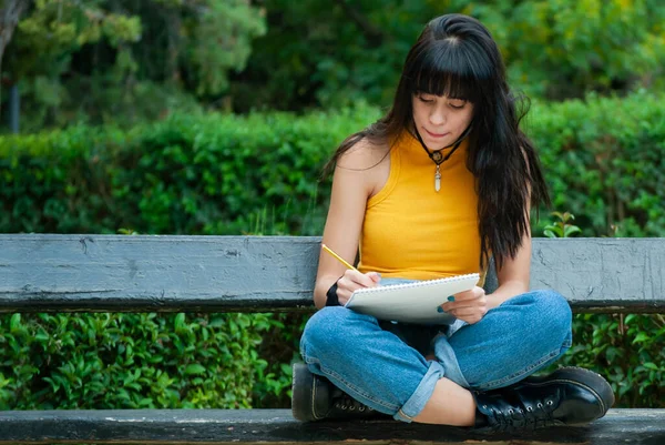 Sarı giyinmiş ve kot pantolon giyen genç bir kız bir not defterine yazarken ya da çizerken bankta oturuyor, sonbahar yeşili ve sarı renkler parkı.