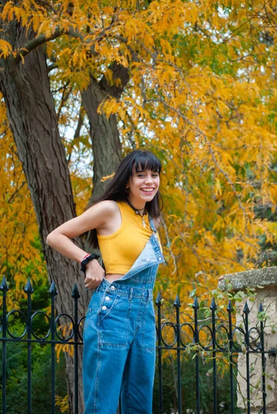Sarı giyinmiş ve kot pantolon giymiş genç bir kız sonbahar yeşili ve sarı renkleri olan bir parkta gülümsüyor..