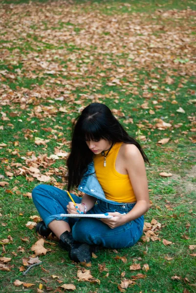 Sarı giyinmiş ve kot pantolon giyen genç bir kız not defterine yazarken ya da çizerken, sonbahar yeşili ve sarı renkler parkında.