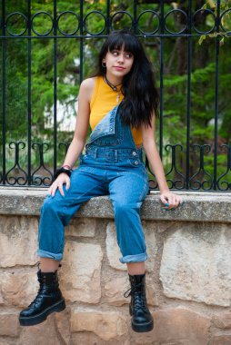 Sarı giyinmiş ve kot pantolon giymiş genç bir kız sonbahar yeşili ve sarı renkleri olan bir parkta taş bir duvarda oturuyor.