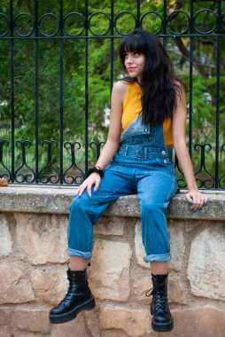 Sarı giyinmiş ve kot pantolon giymiş genç bir kız sonbahar yeşili ve sarı renkleri olan bir parkta taş bir duvarda oturuyor.