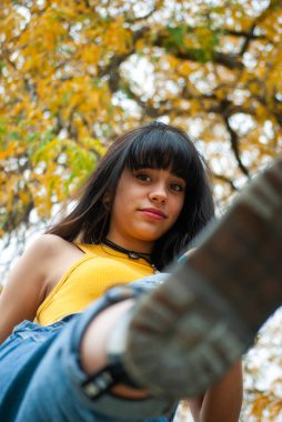 Sarı giyinmiş ve kot tulumlu genç bir kız sarı ve yeşil sonbahar dallarını ve yapraklarını gösteren yakın plan bir çekimde öne doğru tekmeliyor.