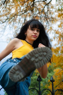 Sarı giyinmiş ve kot tulumlu genç bir kız sarı ve yeşil sonbahar dallarını ve yapraklarını gösteren yakın plan bir çekimde öne doğru tekmeliyor.