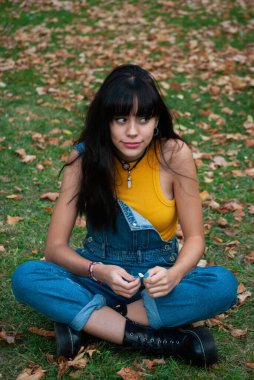 Sarı giyinmiş, kot pantolon giymiş genç bir kız sonbahar yeşili ve sarı renkli bir parkta papatyayla oynarken oturuyor.