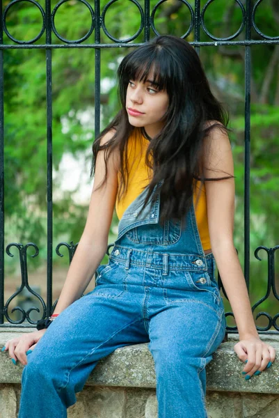 Sarı giyinmiş ve kot pantolon giymiş genç bir kız sonbahar yeşili ve sarı renkleri olan bir parkta taş bir duvarda oturuyor.