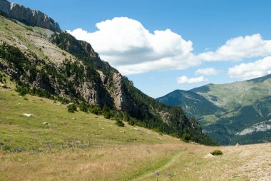 Dağlarla çevrili yeşil vadi ve bulutlu açık mavi bir gökyüzü.