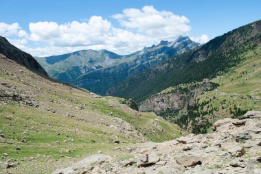 Dağlarla çevrili yeşil vadi ve bulutlu açık mavi bir gökyüzü.