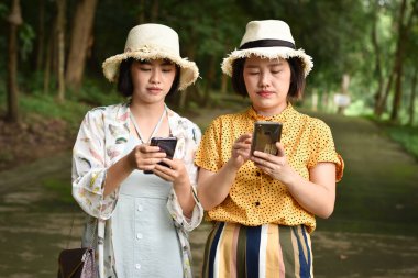 İki Asyalı kız arkadaş açık hava parkında internette gezerken cep telefonu kullanıyor. Jenerasyon dijital iletişim kavramı.