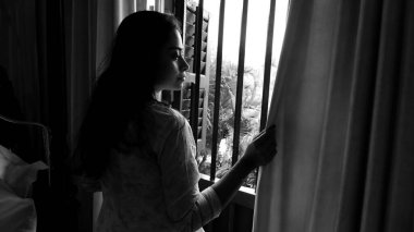 Kolkata, Hindistan, tarih 03.12.2021. Pencere perdesi deliğinden dışarı bakan bir kadının siyah ve beyaz görüntüsü..