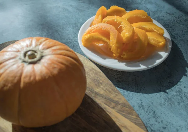 Stock image A small ripe pumpkin on a wooden chopping board next to sliced pumpkin on a white plate on a dark background. Traditional autumn food in the rays of the warm autumn sun. Focus on sliced pumpkin