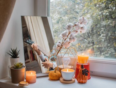 Kız tatil için dekore edilmiş pencere pervazında odasında oturuyor ve ukulele çalıyor. Yansıması aynada, yanan mumlar, çiçekler ve balkabağı heykelciklerinin yanında görülebilir. Tatil için ev dekorasyonu, Şükran Günü, Cadılar Bayramı, doğum günü. 