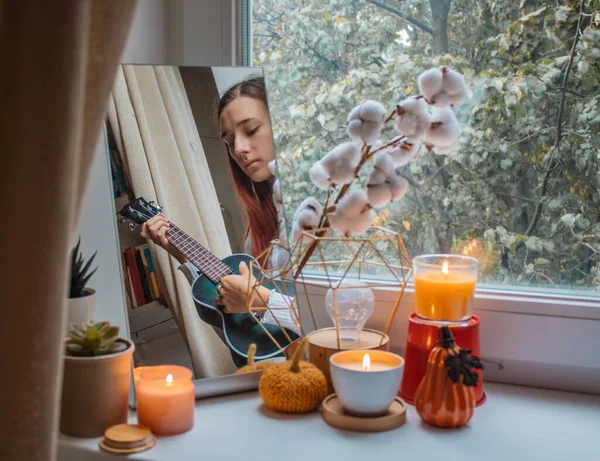 Kız tatil için dekore edilmiş pencere pervazında odasında oturuyor ve ukulele çalıyor. Yansıması aynada, yanan mumlar, çiçekler ve balkabağı heykelciklerinin yanında görülebilir. Tatil için ev dekorasyonu, Şükran Günü, Cadılar Bayramı, doğum günü. 