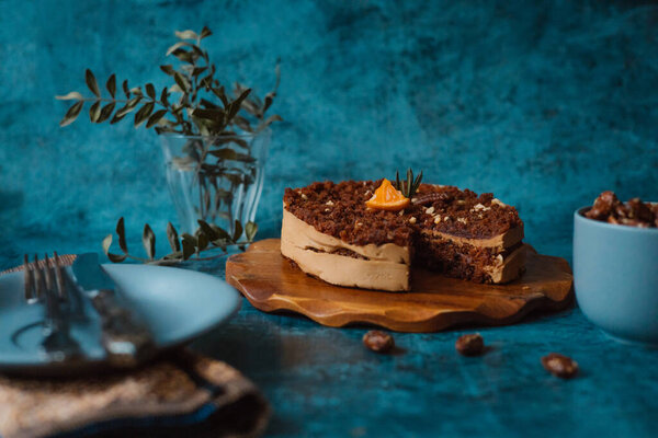 Homemade cake with chocolate and cream on round wooden board next to a plate with a fork and knife and a mug filled with nuts on dark background. Home baking