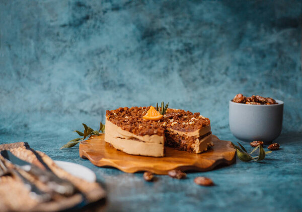 Homemade cake with chocolate and cream on round wooden board next to a plate with a fork and knife and a mug filled with nuts on dark background. Home baking