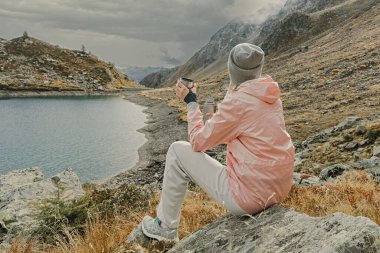 Genç bir kadın turist, dağlarda yüksek bir dağ gölü manzarası izlerken termostan sıcak çay ya da kahve içer..