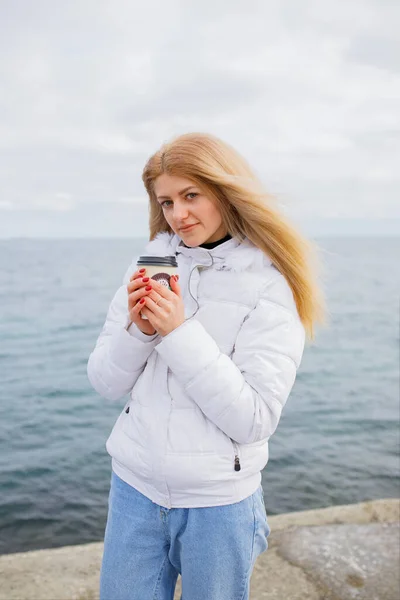 若いブロンドの女性が海辺の近くに立ってコーヒーを飲むと 寒い冬の私達の側は 低温です 白いジャケットとジーンズ 赤い爪 長い髪 カメラを見て — ストック写真