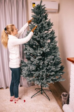  Beyaz örülü süveter ve kot pantolonlu genç sarışın kadın toplar ve oyuncaklarla süslüyor, yapay yeni yıl ağacını ve akşamları evini ateşe veriyor. Noel tatili kavramı. Yaklaşan 2022 Yeni Yıl