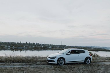 Siyah güneş gözlüklü, beyaz tişörtlü yakışıklı adam modern beyaz spor arabasında oturur ve gün batımını seyreder. Riverside yakınlarında şehir dışında bir hafta sonu. Koyu renkli fotoğraf