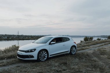 Siyah güneş gözlüklü, beyaz tişörtlü yakışıklı adam modern beyaz spor arabasında oturur ve gün batımını seyreder. Riverside yakınlarında şehir dışında bir hafta sonu. Koyu renkli fotoğraf