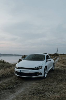 Siyah güneş gözlüklü, beyaz tişörtlü yakışıklı adam modern beyaz spor arabasında oturur ve gün batımını seyreder. Riverside yakınlarında şehir dışında bir hafta sonu. Koyu renkli fotoğraf