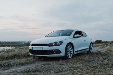 Siyah güneş gözlüklü, beyaz tişörtlü yakışıklı adam modern beyaz spor arabasında oturur ve gün batımını seyreder. Riverside yakınlarında şehir dışında bir hafta sonu. Koyu renkli fotoğraf