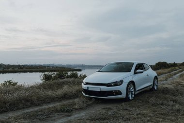 Siyah güneş gözlüklü, beyaz tişörtlü yakışıklı adam modern beyaz spor arabasında oturur ve gün batımını seyreder. Riverside yakınlarında şehir dışında bir hafta sonu. Koyu renkli fotoğraf