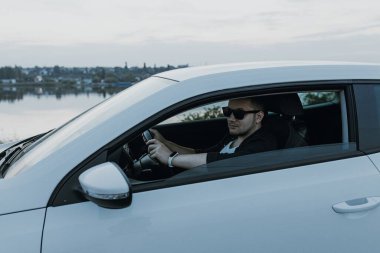Siyah güneş gözlüklü, beyaz tişörtlü yakışıklı adam modern beyaz spor arabasında oturur ve gün batımını seyreder. Riverside yakınlarında şehir dışında bir hafta sonu. Koyu renkli fotoğraf