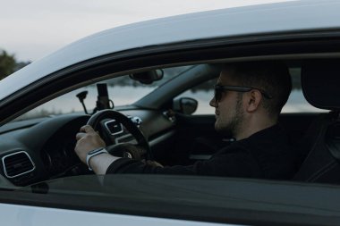 Siyah güneş gözlüklü, beyaz tişörtlü yakışıklı adam modern beyaz spor arabasında oturur ve gün batımını seyreder. Riverside yakınlarında şehir dışında bir hafta sonu. Koyu renkli fotoğraf