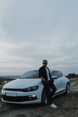 Siyah tişörtlü, kot pantolonlu ve spor ayakkabılı yakışıklı bir adam modern beyaz spor arabanın yanında poz veriyor ve gün batımını seyrediyor. Riverside yakınlarında şehir dışında bir hafta sonu. Koyu renkli fotoğraf