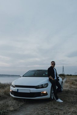 Siyah tişörtlü, kot pantolonlu ve spor ayakkabılı yakışıklı bir adam modern beyaz spor arabanın yanında poz veriyor ve gün batımını seyrediyor. Riverside yakınlarında şehir dışında bir hafta sonu. Koyu renkli fotoğraf